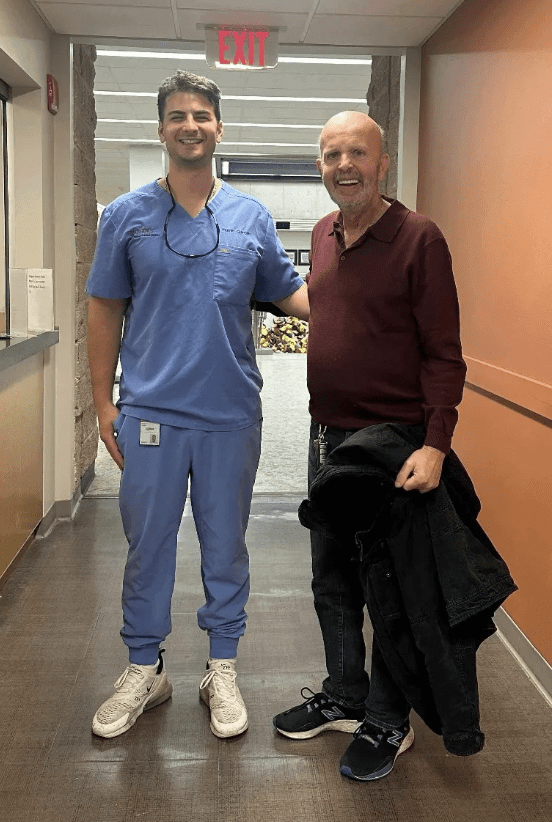 Two men posing together in a hallway, one in scrubs.